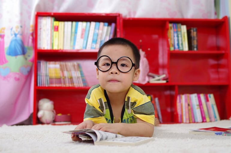 Young child wearing glasses lying on the floor in front of a red bookcase, reading a book with focus