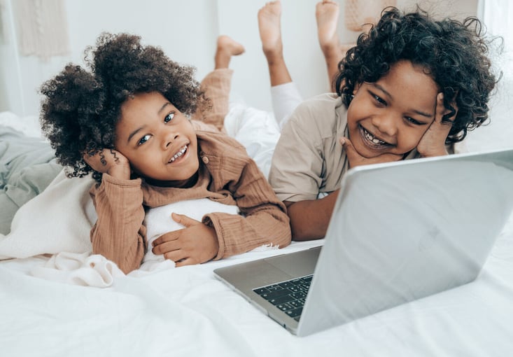 Mother and child lying on a bed, smiling and watching something on a laptop together in a cozy home setting