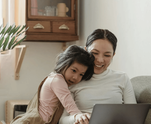 Mother working on a laptop at home with her daughter snuggled beside her, illustrating work-life balance and flexible care