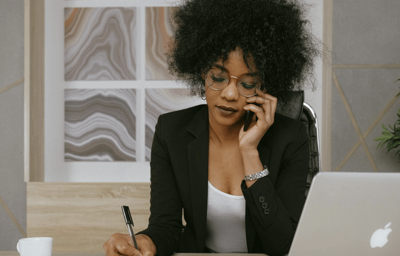 A focused Black woman in professional attire talks on a phone while writing at a desk.