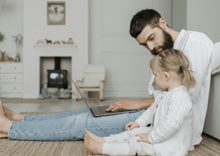 A man with a beard and a young child are sitting on the floor, looking at a laptop. The man has a focused expression, and the child is looking up at the screen. They are in a well-lit room with light-colored walls and a fireplace in the background.