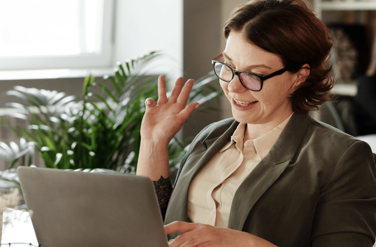 Woman sitting at a desk waving at her laptop while smiling, engaged in a video call