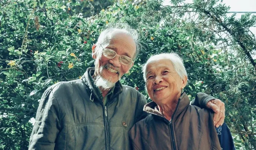Elderly couple standing outdoors with arms around each other, smiling and enjoying a moment together