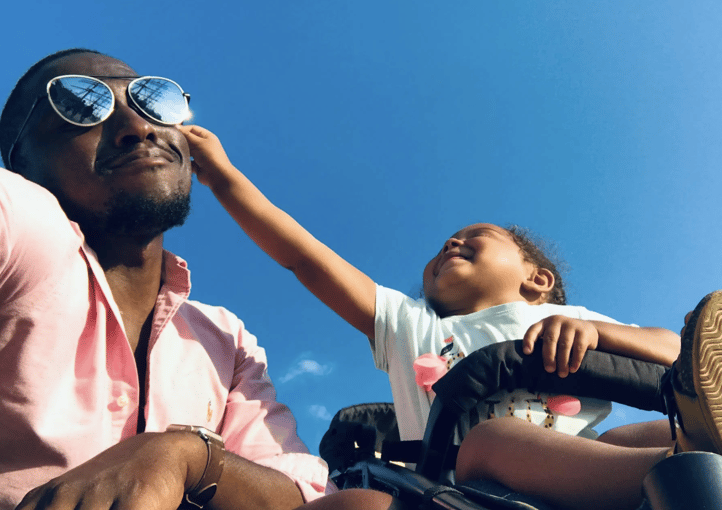 Father wearing sunglasses pushing his child in a stroller, child reaching up toward him against a clear blue sky
