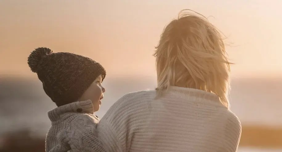 Parent and child wearing warm clothes standing together, looking out at a sunset landscape