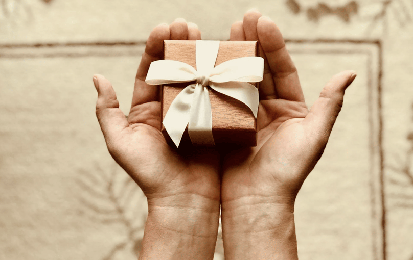 Hands holding a small gift box wrapped in brown paper with a white ribbon against a textured background