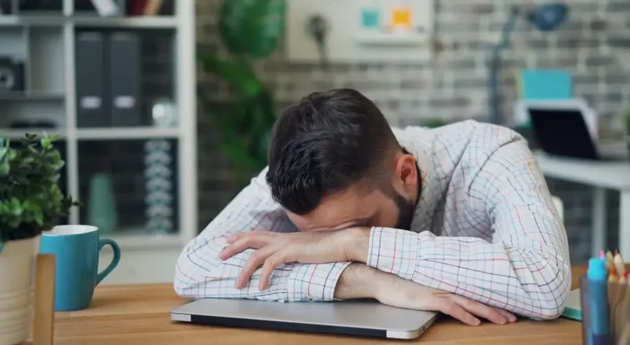 Stressed employee resting his head on folded arms at a desk surrounded by work materials