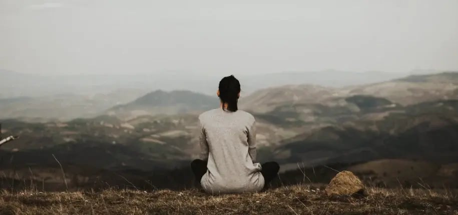Person sitting cross-legged on a hilltop, facing a panoramic mountain view, meditating in solitude