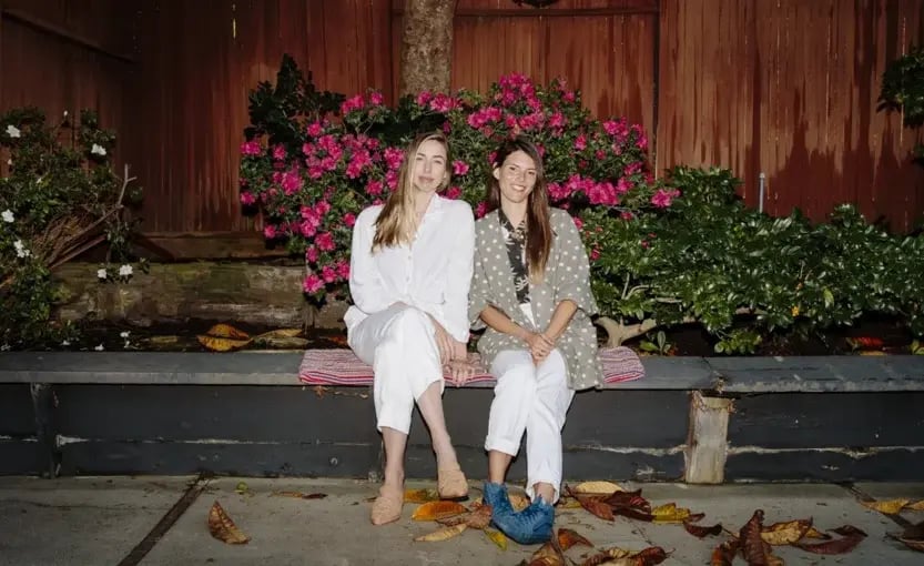 Two women sitting on a bench in front of a building with flowers, smiling and enjoying a relaxed moment together