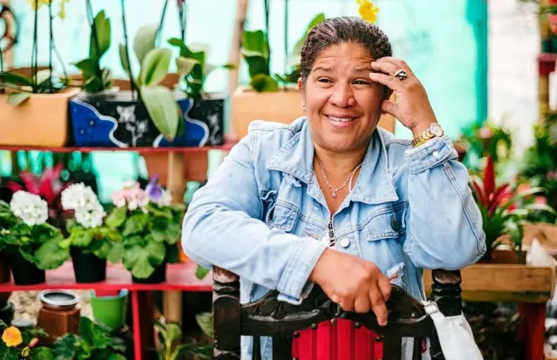 A smiling older woman with short, dark hair is leaning on a walking aid, looking directly at the camera. She is wearing a denim jacket and a red shirt, with various plants and flowers in the background.