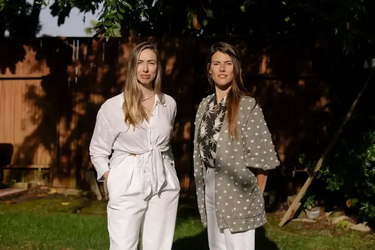 Two women standing side by side outdoors, looking confidently at the camera with a garden backdrop