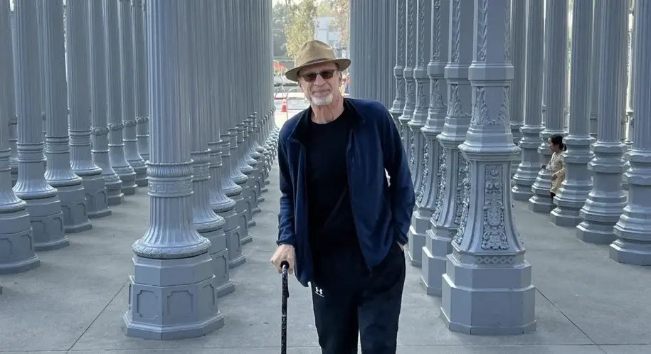 Man walking with a cane through an outdoor art installation of tall lamp posts, smiling during his recovery