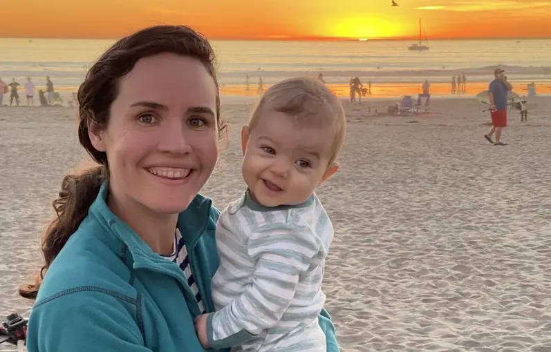 Smiling mother holding her baby on a beach at sunset, capturing a joyful moment during maternity leave