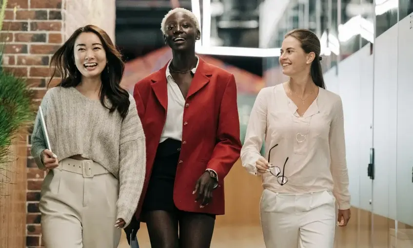 Three smiling women of diverse backgrounds walk together in an office building.