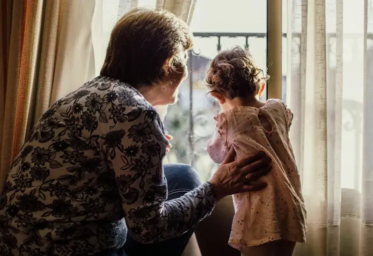 Elderly person sitting with a young girl by the window, sharing a warm moment that reflects intergenerational caregiving