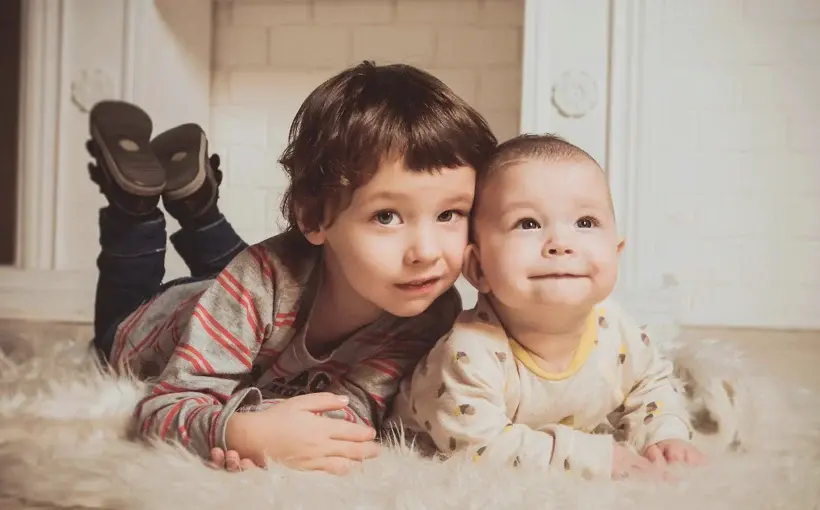 Two young siblings lying on a fluffy rug indoors, smiling and enjoying a cozy moment together
