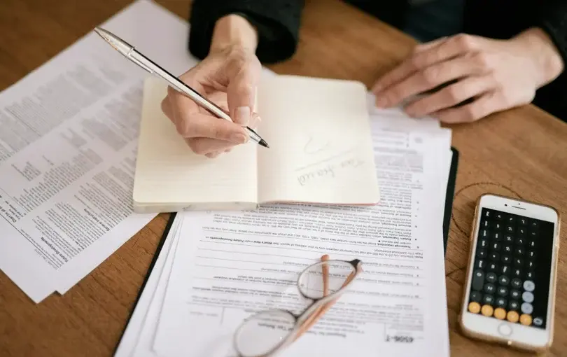 Person writing notes in a notebook next to financial documents, glasses, and a calculator on a wooden desk