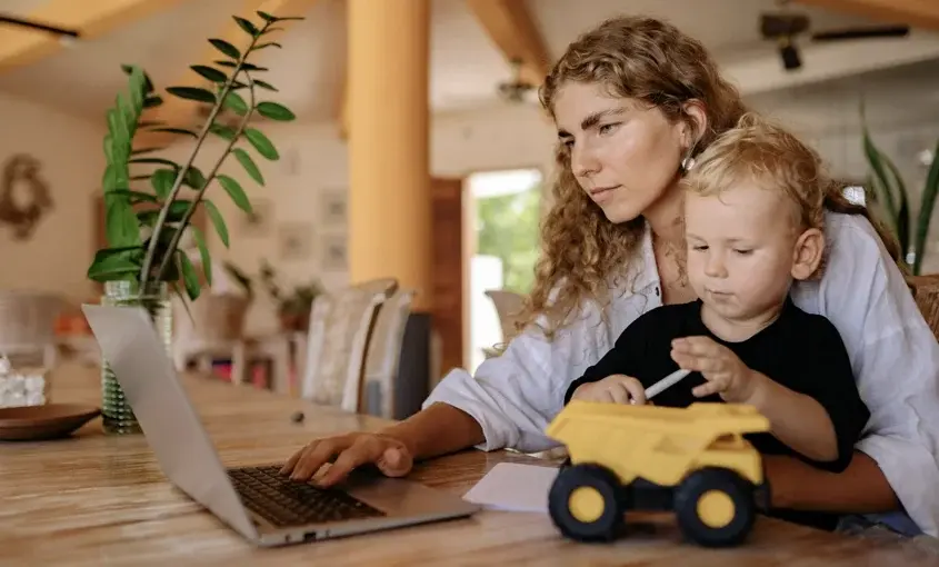 Mother working on a laptop at home while holding her young child with a toy truck on the table