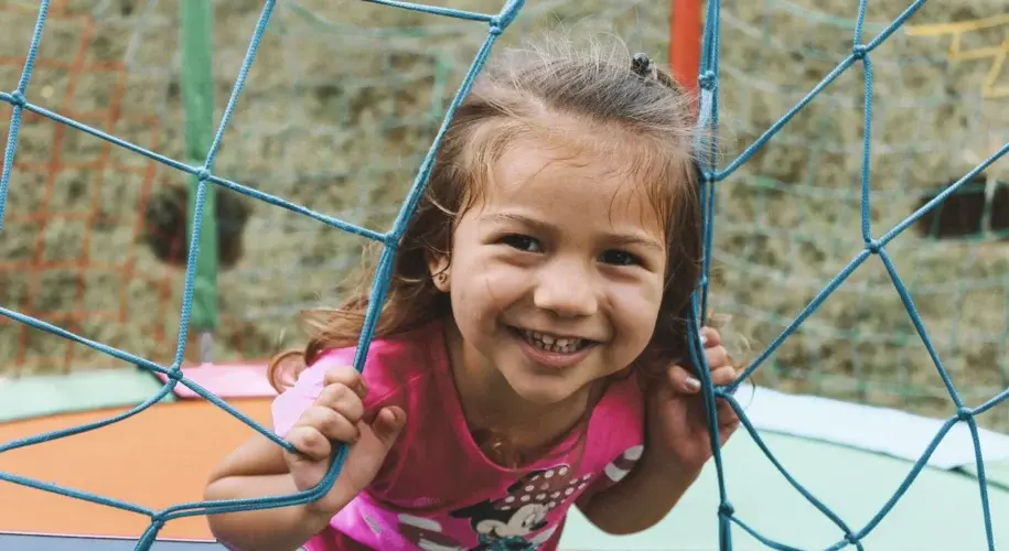 A young girl smiles cheerfully, looking through a metal fence on a sunny day.