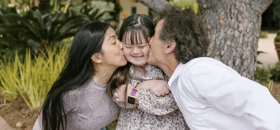 Young girl with Down syndrome smiling as she is kissed on both cheeks by two adults, capturing a moment of love and inclusion