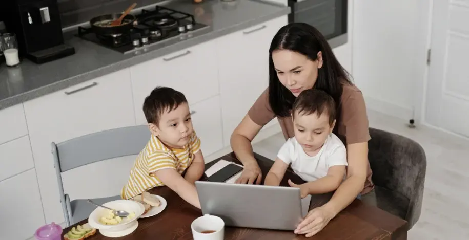 A woman is seated at a kitchen table, looking at a laptop, with two young children beside her, also looking at the screen. One child is on a chair next to her, and the other is on her lap.