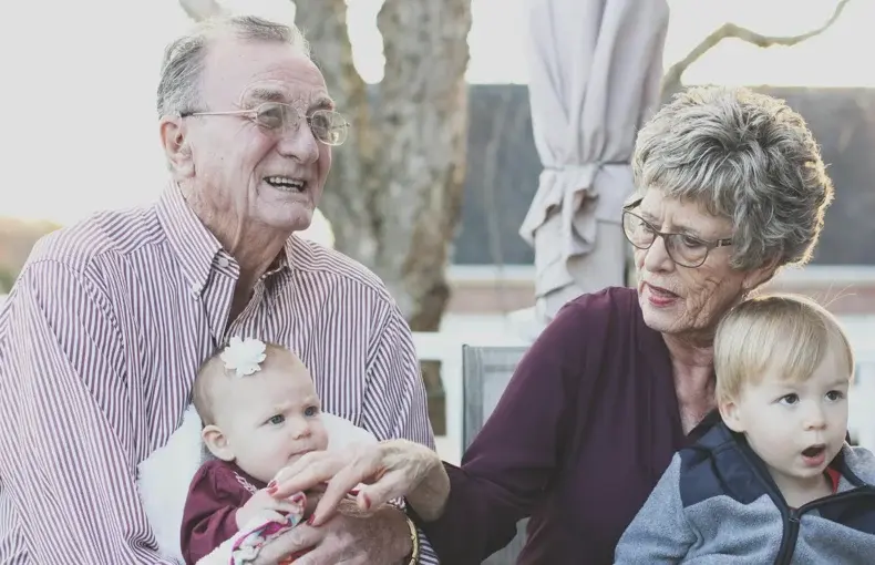 A warm, candid photo shows an elderly man holding a baby, while an elderly woman sits beside him looking at another infant.