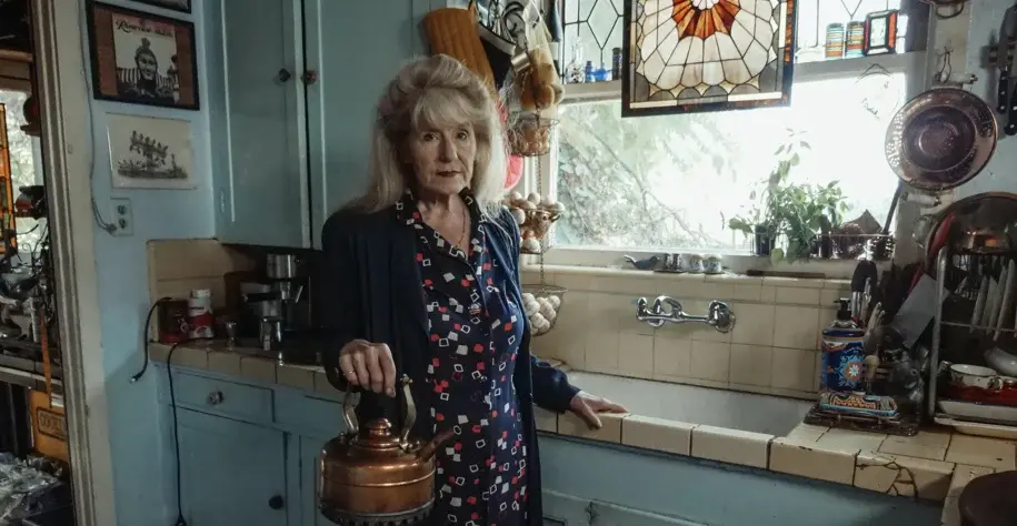 Elderly woman standing in a cozy kitchen holding a kettle, surrounded by vintage decor and household items
