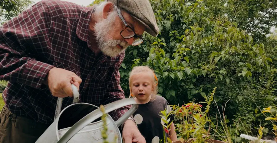 Older man watering plants with a young child in a garden, sharing a moment of intergenerational bonding