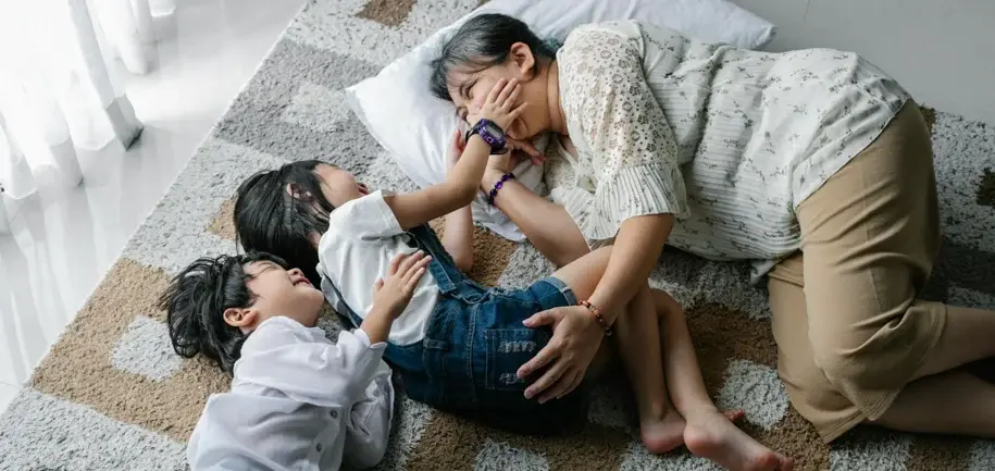 An adult, possibly a caregiver, is lying on the floor on a rug, interacting with two children. One child is lying on their stomach, and the other is lying on their back. The adult is smiling and appears to be playing with the children.