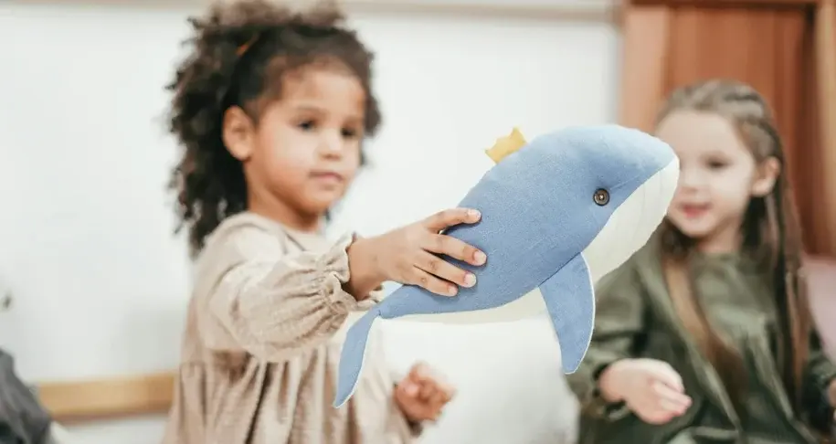 Young girl holding a stuffed toy dolphin in a classroom setting, engaging in a playful and emotionally expressive activity