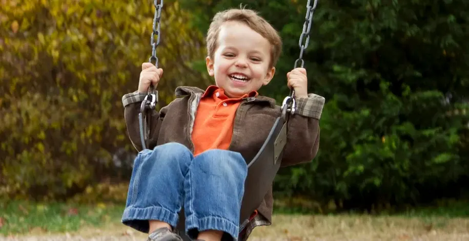 A young boy with a wide smile is on a swing set, looking towards the camera. He is wearing a brown jacket over an orange shirt and blue jeans, with autumn trees blurred in the background.