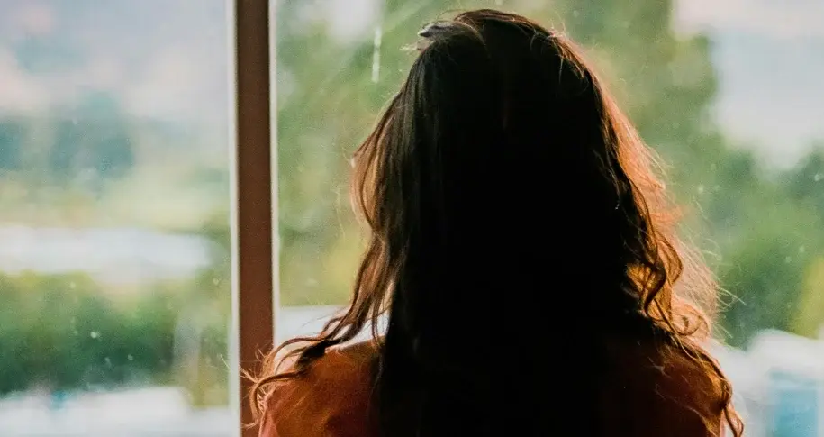 Person with long hair sitting alone and looking out a window at a blurred green landscape, conveying reflection and solitude