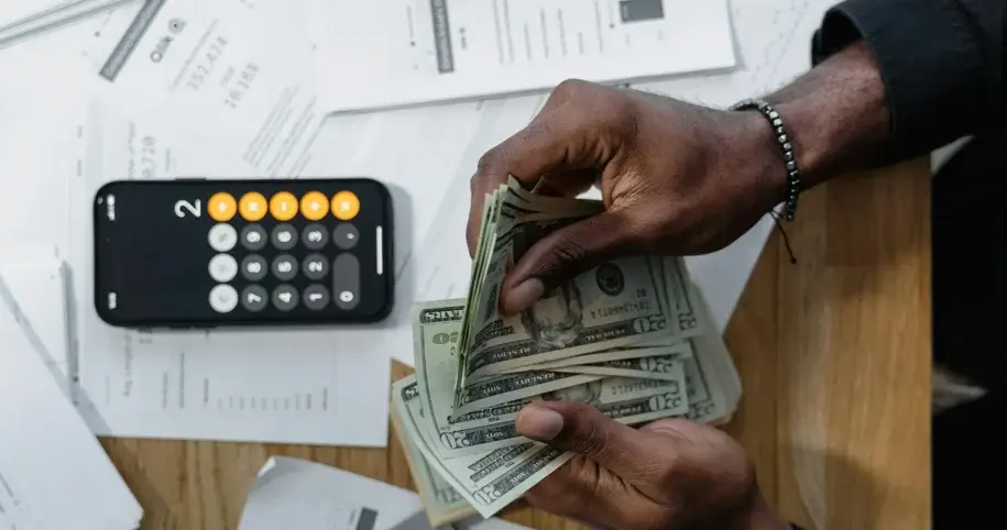 A person's hands counting a stack of US dollar bills next to a calculator.