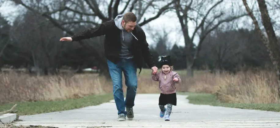 Father walking alongside his toddler daughter on an outdoor path, encouraging and guiding her as they move together