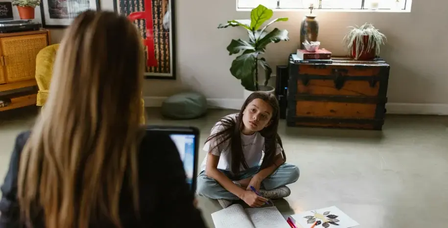 A child sitting on the floor looking at a person whose back is to the camera, in a cozy indoor setting.