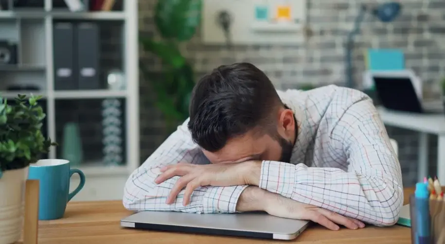 Stressed employee resting his head on folded arms at a desk surrounded by work materials