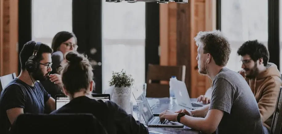 A diverse group of young adults are working together in a modern office setting. They are seated around a table with laptops and headphones, engaged in collaborative work or discussions.