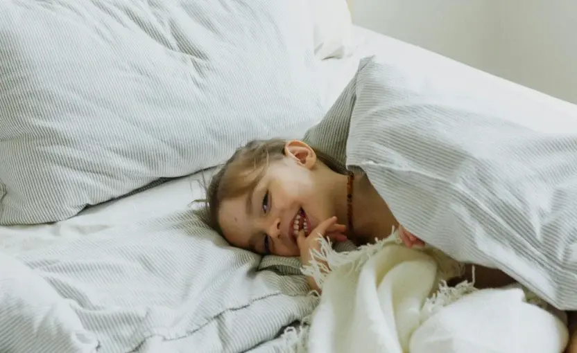 Child tucked under a white blanket, peeking out from under pillows in a cozy bedroom