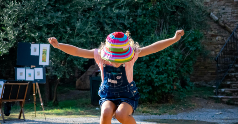 Child wearing a colorful striped sun hat jumping outdoors with arms outstretched