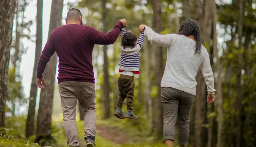 Two adults walking through a forest trail while holding a child's hands and lifting them playfully