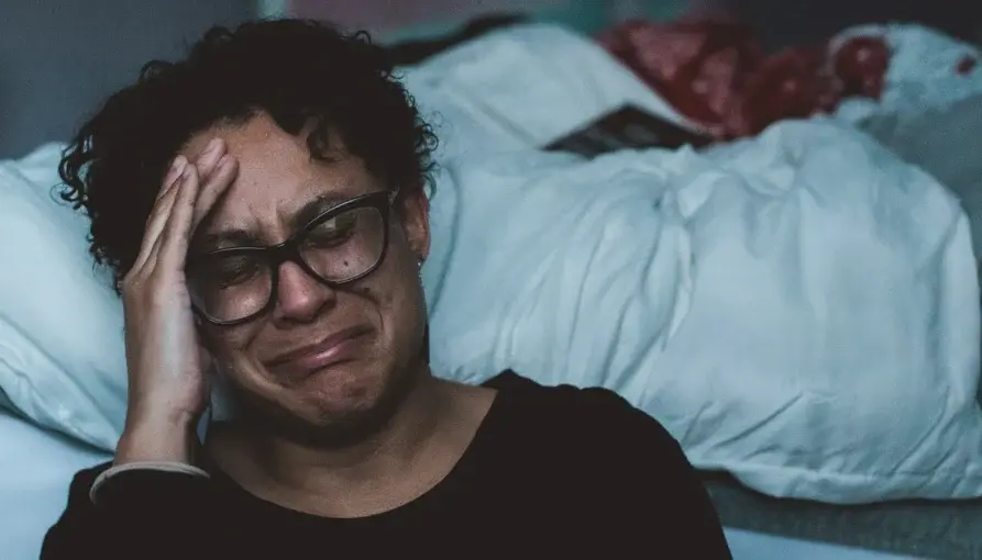 Stressed caregiver sitting on a bed with a hand on their forehead, appearing overwhelmed and tearful