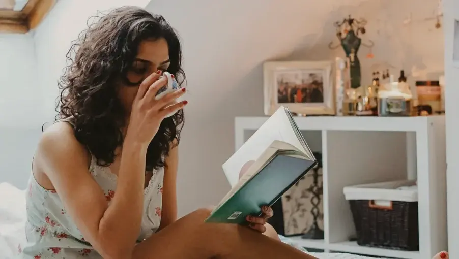 A person with curly hair is sitting on a bed or couch, holding a clear glass of water and reading an open book. They are wearing a light-colored, possibly floral, top. In the background, there are various items on a shelf or dresser.