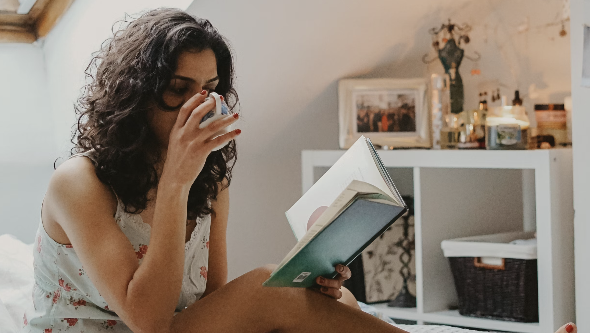 a woman, sitting in bed, drinking coffee, and reading a book