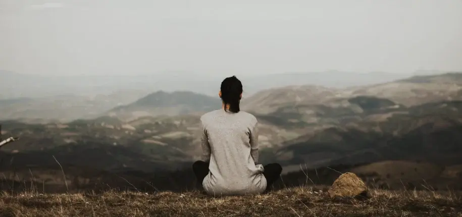 Person sitting cross-legged on a hilltop, facing a panoramic mountain view, meditating in solitude