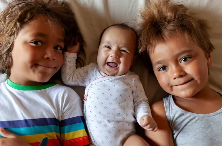 A diverse group of three happy children, including a baby, lying together and smiling.