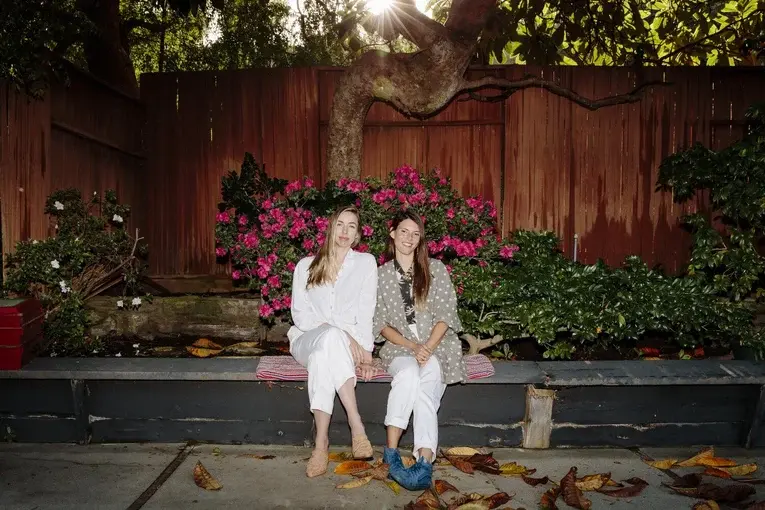 Two women sit side-by-side on a stone bench in front of a wooden fence.